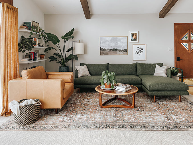 A cozy living room with a green sectional sofa and a tan leather armchair. The room features a wooden coffee table with plants, a patterned rug, and wall art. Shelves with books and decor are on the left, and a door with a window is on the right.