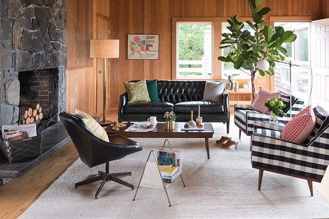 A cozy living room with wood-paneled walls features a stone fireplace with logs stacked inside. The room has a black leather sofa, a black swivel chair, and a checkered armchair. A wooden coffee table holds books and plants, and large windows let in natural light.