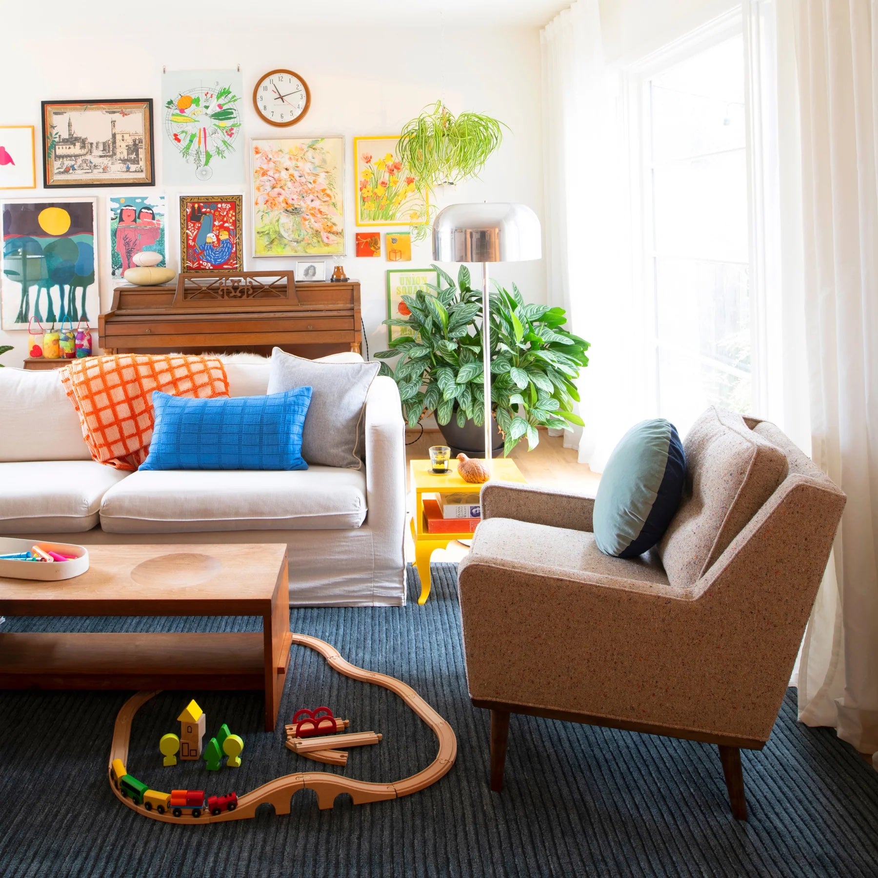 A cozy living room features a white sofa with colorful pillows, adjacent to a beige armchair. A wooden coffee table stands between them. A toy train set is on the floor. The walls are adorned with various artworks, and green plants add to the room's lively atmosphere.