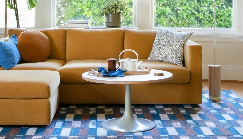 A cozy mustard-colored sectional sofa with a variety of pillows, including round and patterned ones, is against a backdrop of large windows with greenery. A round coffee table in front displays a tea set, tray, and napkin. The floor features a checkered rug.