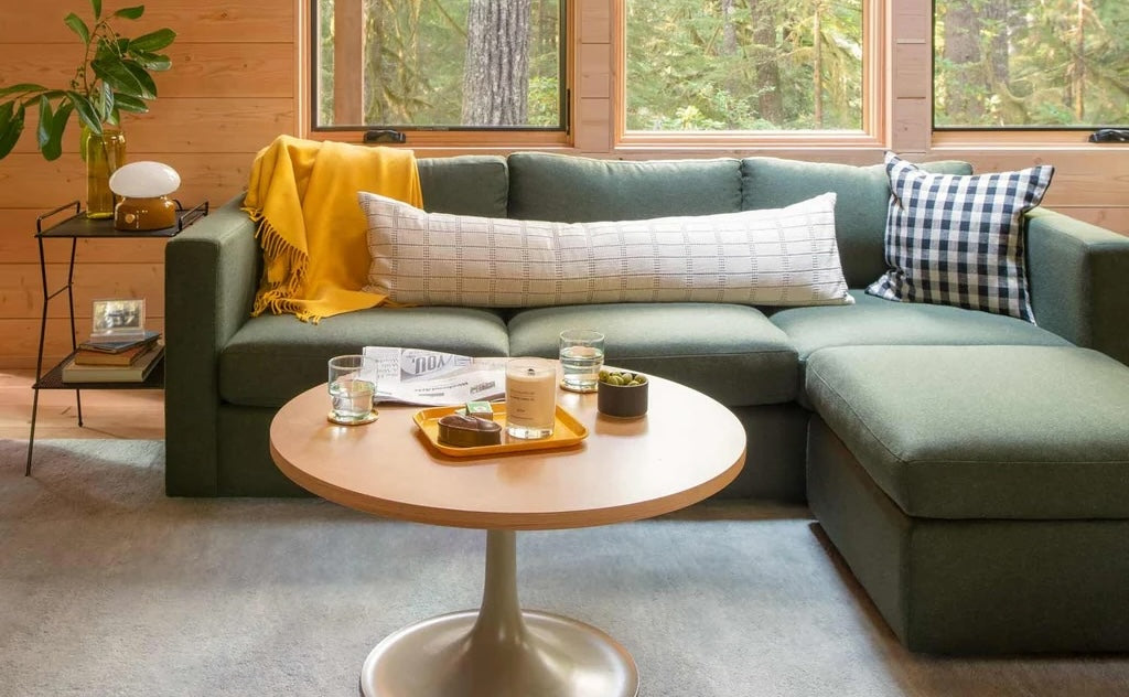 A cozy living room with a green sectional sofa adorned with a yellow throw blanket and patterned pillows. A round wooden coffee table in front holds drinks, a candle, and a tray. Large windows offer a view of lush greenery outside, enhancing the room's natural ambiance.