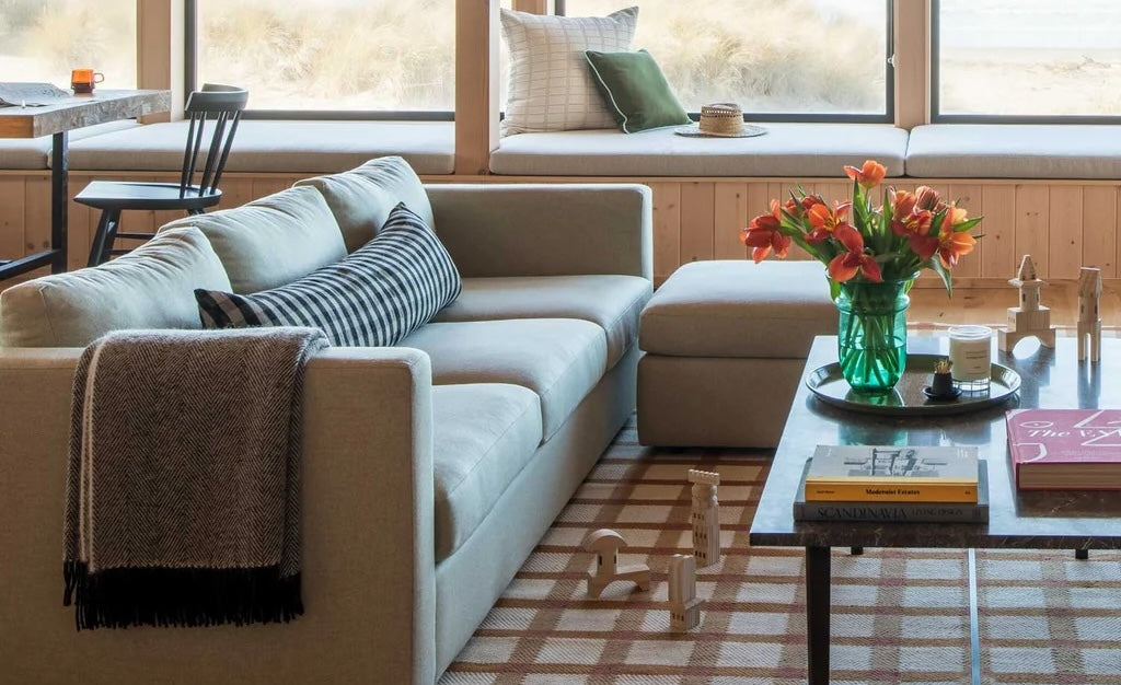 A cozy living room with a light beige L-shaped sofa adorned with a striped cushion and a throw blanket. A coffee table with books and a vase of orange flowers stands in front, while a window bench seat overlooks an outside view. The decor is minimal and inviting.