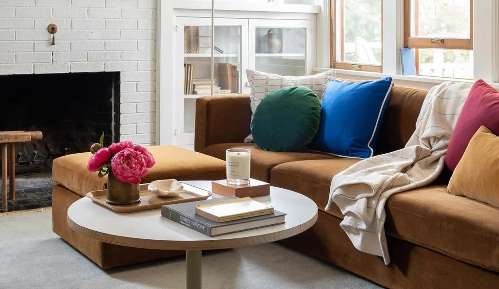 A cozy living room with a brown sectional sofa adorned with various colorful throw pillows and a blanket. A round white coffee table at the center holds books, a candle, and a bowl with fresh pink flowers. A white brick fireplace and windows are in the background.