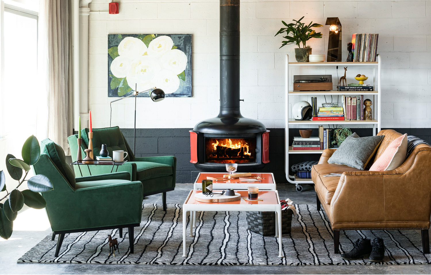 Stylish living room with a cozy fireplace, green armchairs, a brown leather sofa, a patterned rug, and a white coffee table. A bookshelf with plants and decor items stands against the wall. A large floral painting hangs above the fireplace. Sunlight filters in from the left.