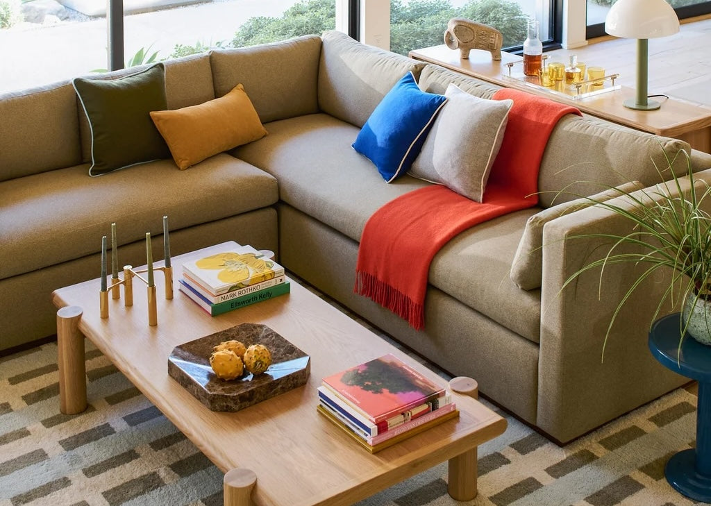 A cozy living room featuring a large beige sectional sofa adorned with colorful throw pillows and a red blanket. A wooden coffee table with candles, books, and decorative items sits on a patterned rug in front of the sofa. Natural light streams in through large windows.
