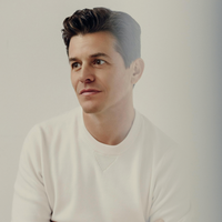 A man in a white sweater gazes to the side with a thoughtful expression. He has short, dark hair styled neatly and is pictured against a plain, light background. The image has a soft, serene feel.