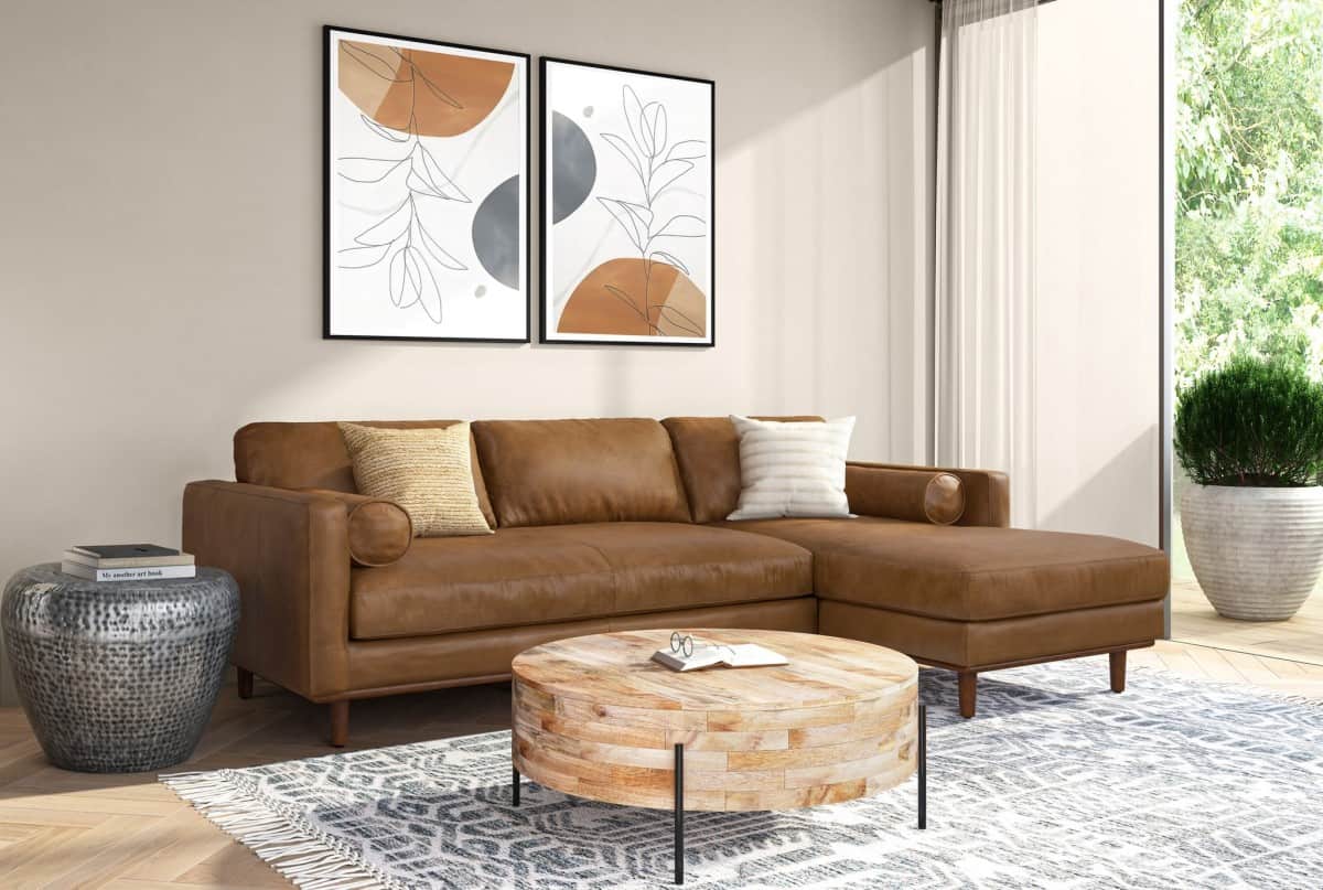 A modern living room features a brown leather sectional sofa with two accent pillows, an abstract art duo on the wall behind, a round wooden coffee table, and a patterned rug. A large window allows natural light to brighten the space, with plants visible outside.