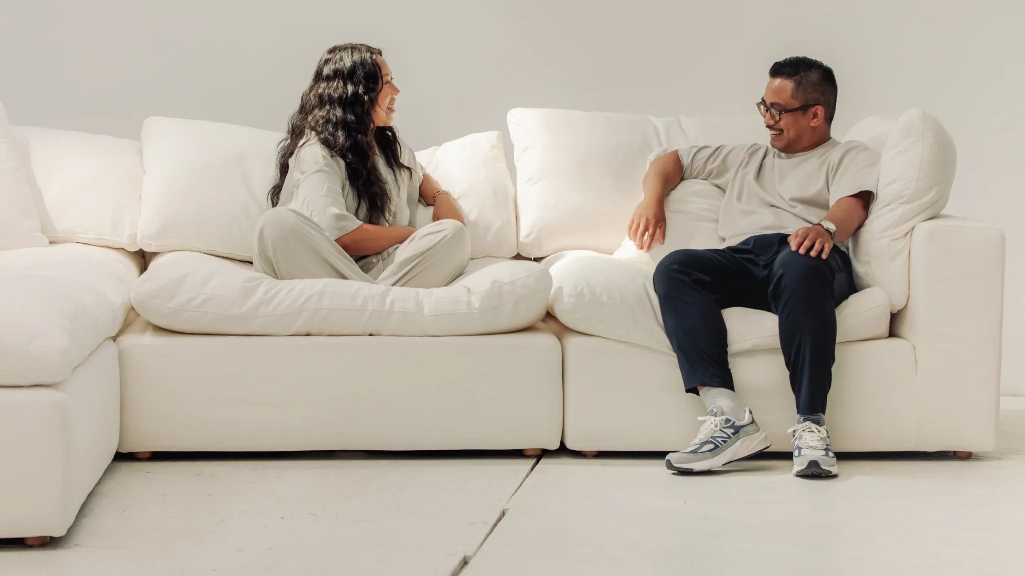 A woman and a man are sitting on a white sectional sofa. Both are smiling and engaged in conversation. They are dressed in casual, light-colored clothing. The background is minimalistic with a white wall and floor, creating a clean and serene atmosphere.