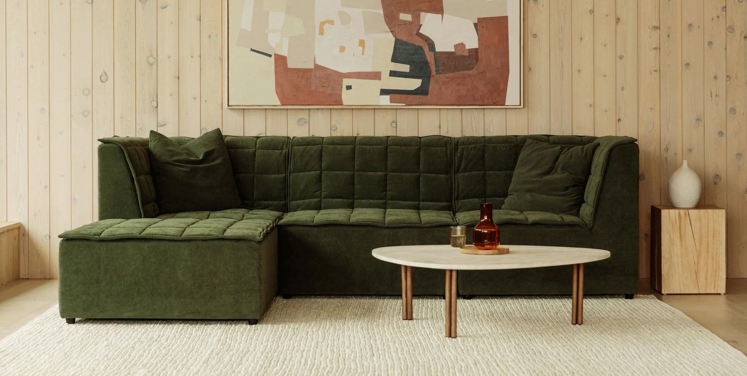 A cozy living room features a plush green sectional sofa with pillows, set against a light wood-paneled wall. Above the sofa hangs an abstract painting in earthy tones. In front of the sofa, a round white coffee table holds a brown glass vase and a candle.