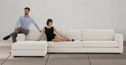 A man wearing a blue shirt and gray pants, along with a woman in a black dress, are seated at opposite ends of the Architect 133" Fabric Sofa Sectional Left in Blanc Boucle, which is placed against a plain wall. The room features a minimalist design with a neutral-toned floor.