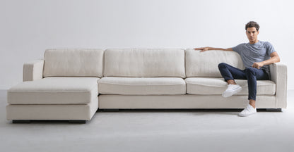 A person in a gray T-shirt and dark pants sits on the Architect 133" Fabric Sofa Sectional Left, Blanc Boucle, which boasts an L-shape design. The room features a minimalist style with plain white walls and a light gray floor.