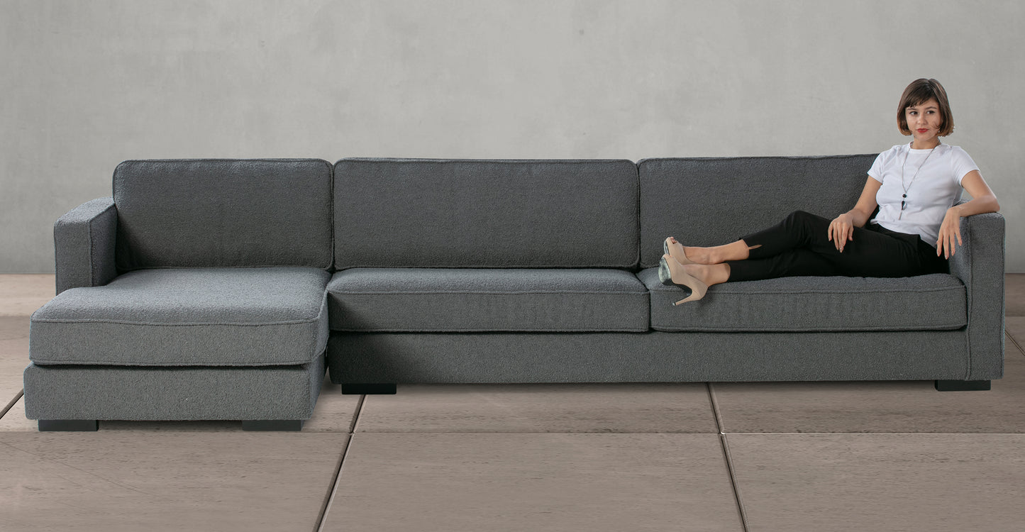 A woman in a white shirt and black pants is reclining on the Architect 133" Fabric Sofa Sectional Left in Blanc Boucle, featuring a modern design. The sofa is set against a plain, light gray wall and flooring, creating a minimalist setting.