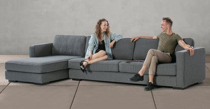 A man and a woman sit on the large Architect 133" Fabric Sofa Sectional Left in Blanc Boucle, facing each other and smiling. The sectional features a chaise lounge on one side. The background showcases a plain, light gray wall and tiled floor.