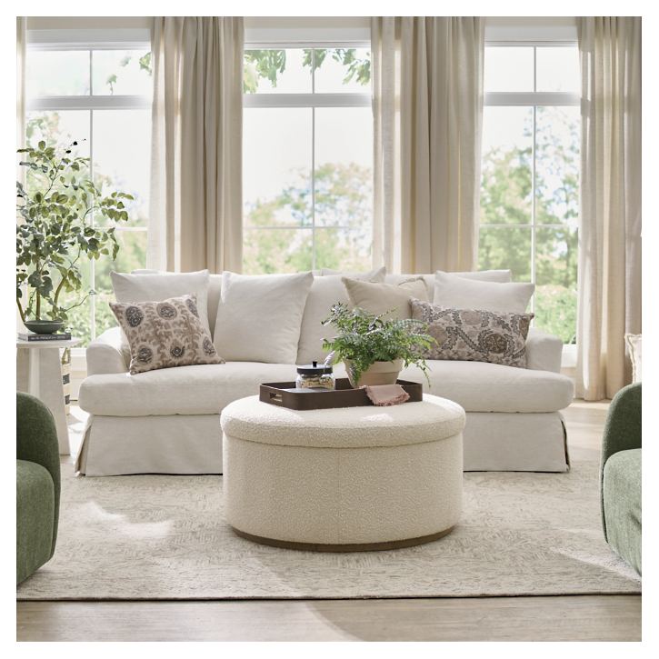 A cozy living room showcases an Ava Slipcovered Sofa adorned with patterned pillows, complemented by a round ottoman holding a tray of decor, large windows draped with flowing curtains, and green armchairs. Sunlight streams in, illuminating the neutral-toned decor and potted plants.