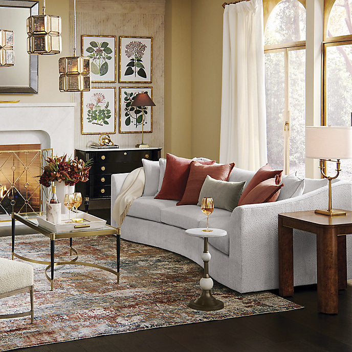 The cozy living room features a Pippa Sofa in white, elegantly adorned with red and gray pillows. A fireplace casts a warm glow, while decorative botanical prints hang above a sleek black console table. The decor is completed with a modern rug and an array of side tables, creating an inviting and stylish atmosphere.