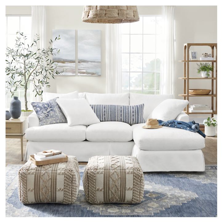 In a cozy living room, the Ava Chaise Slipcovered Sofa is decked out with blue and white patterned pillows. A straw hat and denim jacket rest casually on the sofa. Two textured poufs sit atop a blue patterned rug, while windows and plants contribute to a bright, airy ambiance.