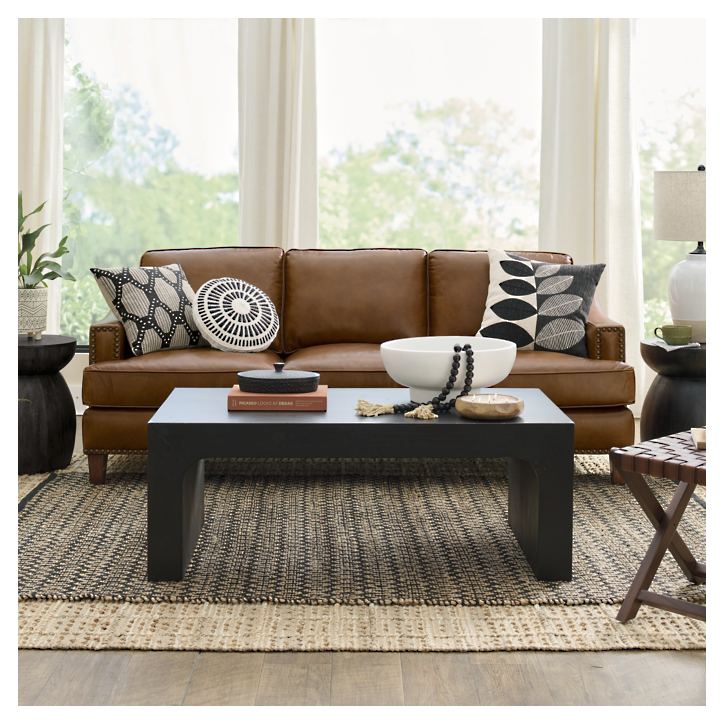 In a stylish living room, the Collins Sofa—a brown leather masterpiece—is adorned with patterned cushions. A dark rectangular coffee table is elegantly placed in front of it, featuring decorative items. A pair of round side tables complement the setup, while large windows draped with light curtains let in natural light. A woven rug adds warmth to the wooden floor beneath.