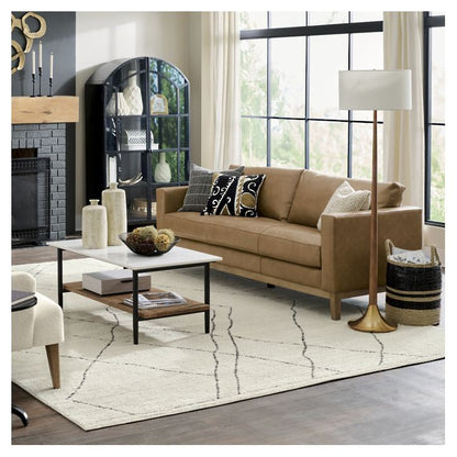 A cozy living room featuring the Capri Sofa in a warm tan color, complemented by a white patterned rug and an elegant floor lamp. In front of the sofa, a wooden coffee table provides a functional centerpiece, while natural light filters through a black-framed window. The space is tastefully decorated with a black cabinet and decorative vases adorning the walls.