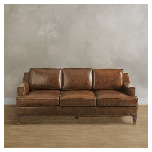 The Collins Sofa, a polished brown leather three-seater, is placed against a neutral, textured gray wall on a wooden floor. It features cushion backrests and armrests, adding a classic and elegant look to the room.