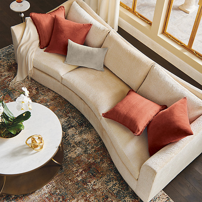 An overhead view of a cozy living room showcases the Pippa Sofa, featuring a cream-colored curved design. The sofa is adorned with red and gray cushions alongside a white throw. A marble-top coffee table displays a gold vase and an orchid, while the floor is accented by a patterned rug.