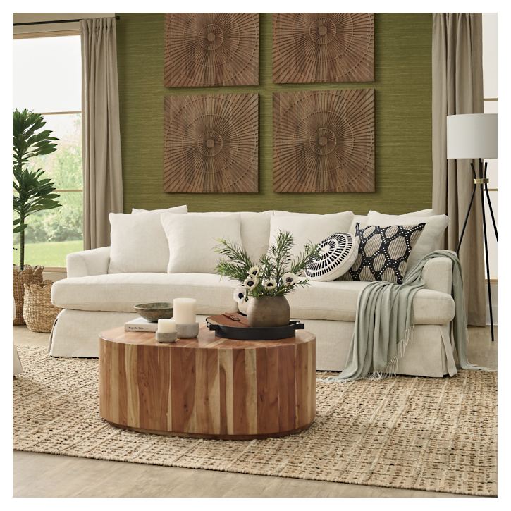 A cozy living room featuring the Ava Slipcovered Sofa adorned with decorative pillows, alongside a wooden coffee table displaying candles and a plant. The green textured wall is enhanced by circular art pieces, with a large window allowing natural light to brighten the space. A potted plant and floor lamp complete the ambiance.