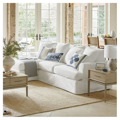 A cozy living room showcases the Ava Chaise Slipcovered Sofa adorned with blue floral pillows, accompanied by a wooden coffee table and a side table topped with a lamp. Large windows flood the room with natural light, while a woven rug contributes texture to the space.