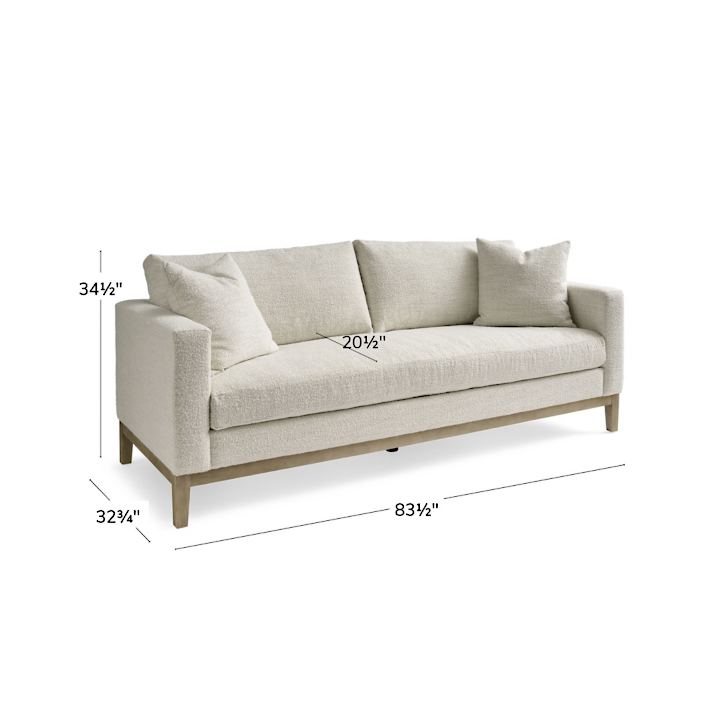 A Capri Sofa featuring a light-colored design with three back cushions and a wooden base. Its dimensions are: height 34.5 inches, depth 32.5 inches, and width 83.5 inches.