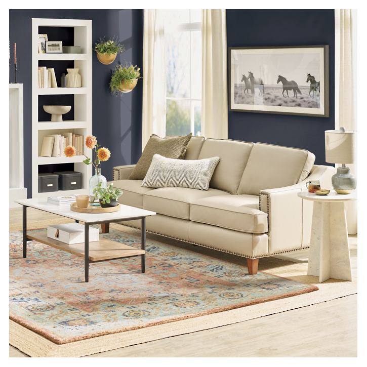 A cozy living room featuring the Collins Sofa, adorned with decorative pillows and a patterned rug. A white shelving unit and wall art enhance the ambiance. A wooden coffee table with potted flowers and a small side table holding a lamp complete the decor as sunlight streams in.