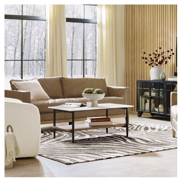 A modern living room featuring the Capri Sofa in beige, accompanied by a coffee table topped with a decorative bowl and a zebra-patterned rug. The space is highlighted by a large window showcasing a snowy view, alongside a striped wood accent wall displaying a cabinet and vase.