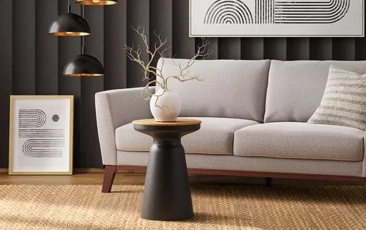 A modern living room features a light grey sofa with a striped pillow, a black side table with a plant vase, and framed abstract geometric art on the wall and floor. The room includes black pendant lights and a woven rug, with a background of dark vertical paneling.