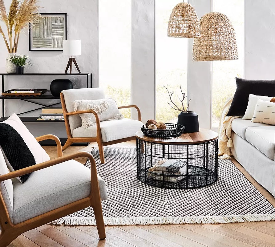 A modern living room with three wooden-framed chairs, a sofa with pillows, a round coffee table with a basket underneath, a black metal shelving unit, and large windows. Pendant lights with wicker shades hang from the ceiling, and a striped rug covers the floor.