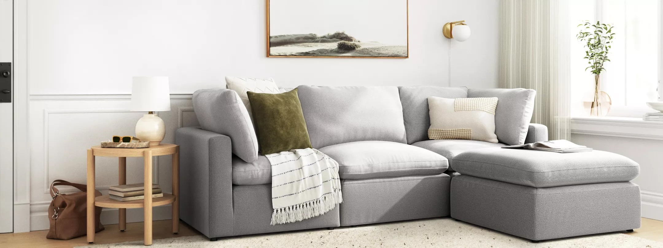A cozy living room corner features a light gray sectional sofa adorned with various throw pillows and a blanket. A round wooden side table holds a lamp and decor items, while a landscape painting and wall-mounted lamp add to the room's ambiance.
