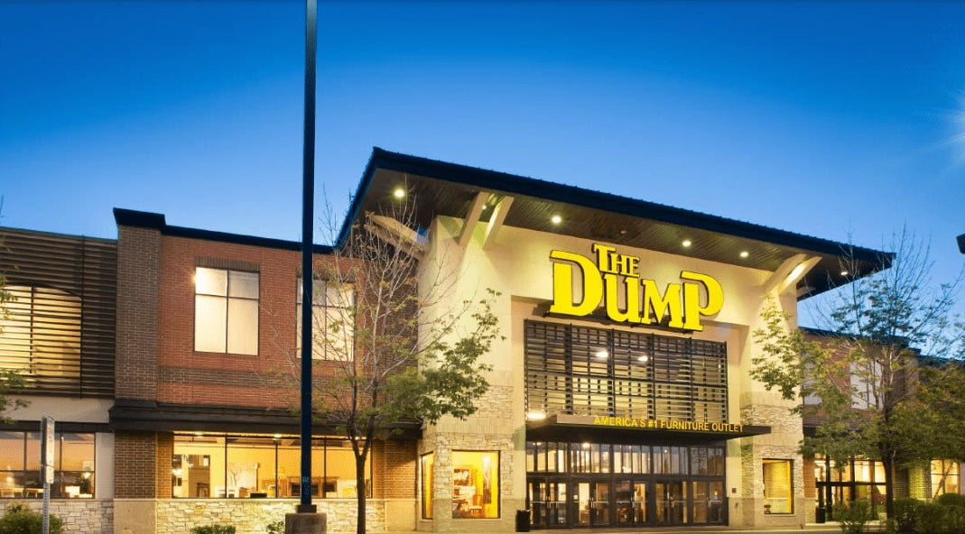A large store named "The Dump" is illuminated by exterior lights at dusk. The building features a mix of brick and stone facade with large windows and a prominent, glowing yellow sign. Trees and a light post are visible in the foreground.
