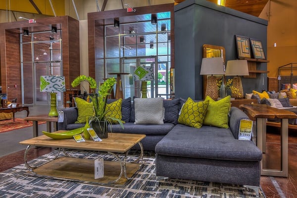 Cozy living room setup in a furniture store featuring a gray sectional sofa adorned with green and gray pillows. Wooden coffee table with decor items, green vase with plants, and table lamp. Modern wall decor and additional furniture pieces are visible in the background.