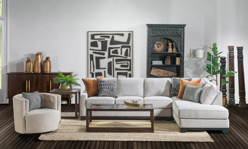 A living room with a white modern sectional sofa adorned with patterned and solid pillows, a glass coffee table, a white cushioned armchair, and a woven rug. A dark wooden cabinet with decor and a tall elegant bookshelf are against the walls, along with some plants.