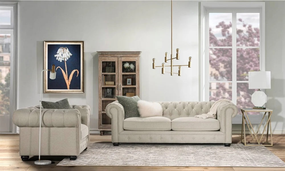 A stylish living room with a beige tufted sofa, matching armchair, and gold-accented furniture. A painting of a white flower hangs on the wall above a wooden cabinet filled with decor items. Large windows show cherry blossoms outside. A modern chandelier hangs above.