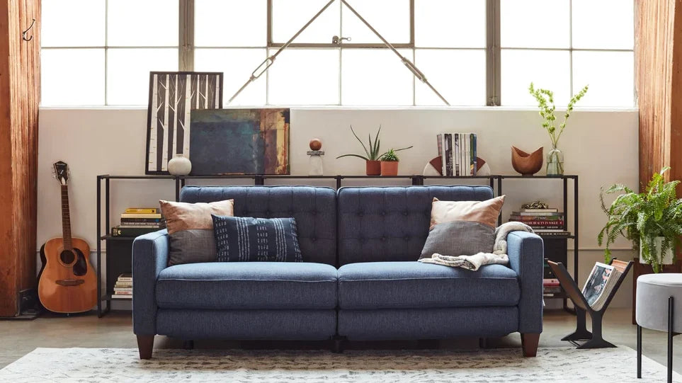 A cozy living room with a blue tufted sofa in the center, adorned with assorted pillows. Behind the sofa is a metal-framed shelf holding books, plants, and decorative items. A guitar rests against the wall, and a large window lets in natural light.