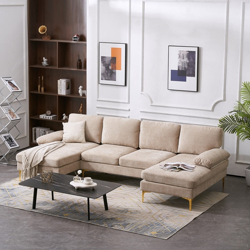 A modern living room features a beige U-shaped 4-seat indoor modular sofa, accompanied by a black coffee table resting on a patterned rug, with two abstract paintings adorning the walls. The space is finished with a shelf showcasing decor items, a potted plant, and an accordion-style magazine rack.