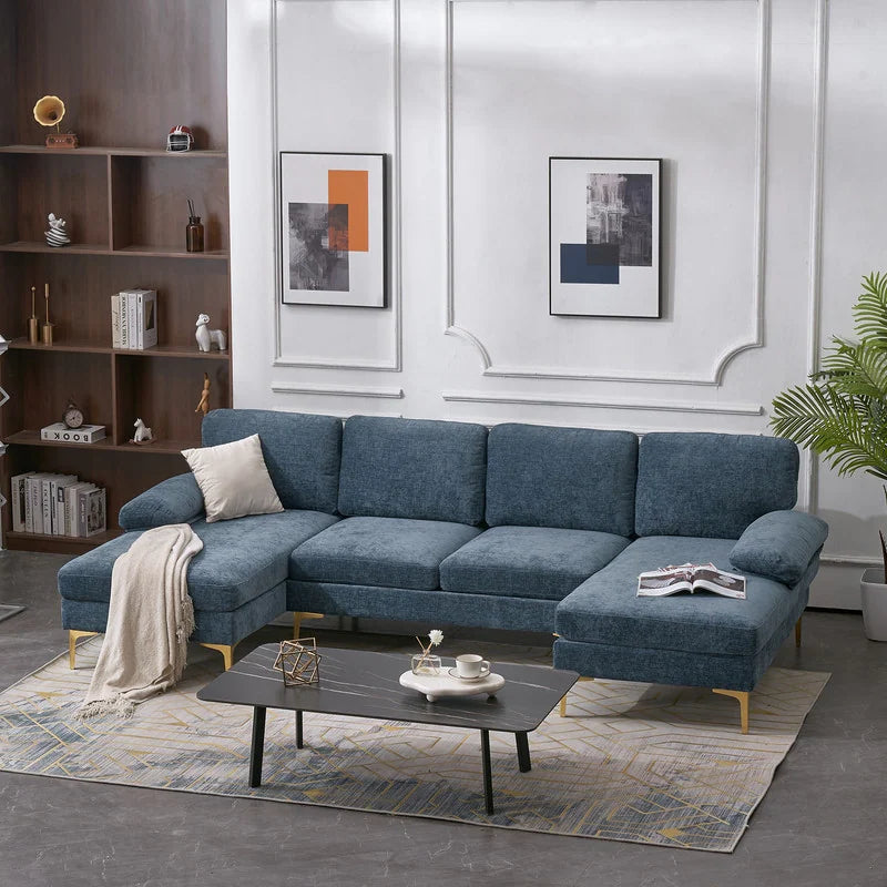 A contemporary living room featuring an elegant U Shaped 4 Seat Indoor Modular Sofa in blue, complemented by a beige throw and cushion. The space includes a sleek black rectangular coffee table, shelves lined with books, two abstract paintings adorning the walls, and a potted plant adding a touch of greenery in the corner.