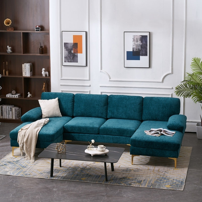 A modern living room featuring a teal U Shaped 4 Seat Indoor Modular Sofa with golden legs, a black coffee table adorned with decorative items, and a patterned rug. Two abstract artworks hang on the white wall, and a wooden shelf filled with books and plants stands in the corner.