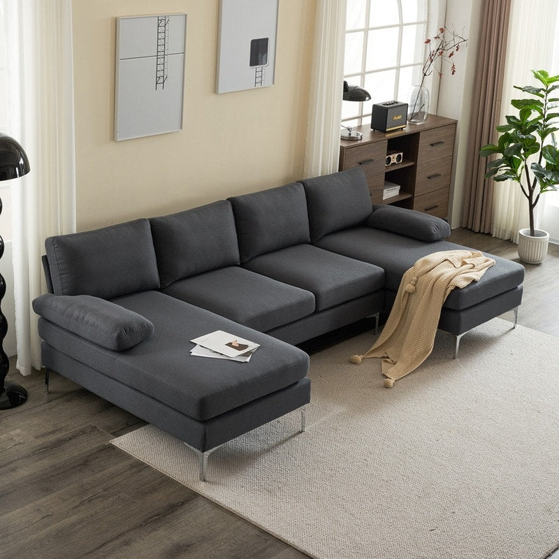 A living room featuring the U Shaped 4 Seat Indoor Modular Sofa in dark gray rests on a light rug. The sofa is adorned with pillows and a beige throw blanket. Two framed artworks grace the wall, and in the background, there's a potted plant alongside a wooden cabinet decorated with various items. Natural light illuminates the space.