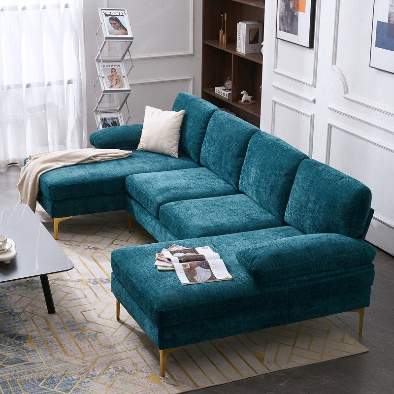 A stylish living room showcases a U Shaped 4 Seat Indoor Modular Sofa in teal velvet, adorned with a cream pillow. Magazines are elegantly placed on the ottoman. The space features a geometric rug, a marble coffee table, a magazine rack, and a wooden bookshelf set against white paneled walls.