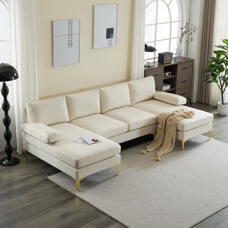 A modern living room featuring an "U Shaped 4 Seat Indoor Modular Sofa" in beige is set on a light grey rug. The space includes a dark floor lamp, a potted plant, wall art, and a wooden cabinet. A beige throw is elegantly draped over the sofa, while large windows flood the area with natural light.