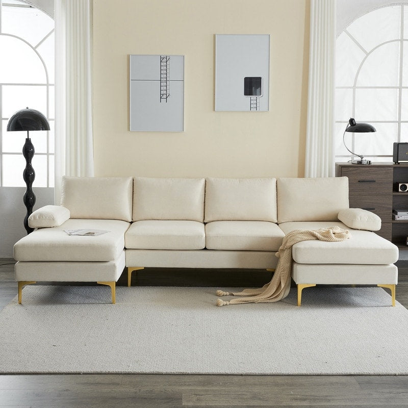 A modern living room featuring the U Shaped 4 Seat Indoor Modular Sofa in cream on a light gray rug. Two minimalist paintings adorn the pale yellow wall, while a black floor lamp and a dark wooden desk with a lamp complete the background. A beige throw blanket is elegantly draped over the sofa.