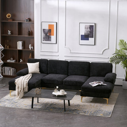 A contemporary living room features the U Shaped 4 Seat Indoor Modular Sofa in black, enhanced by a cream pillow and blanket. A sleek black coffee table showcases decorative items. Two abstract artworks are displayed on the white-paneled wall. Wooden shelves and a green plant add the finishing touches to the space.