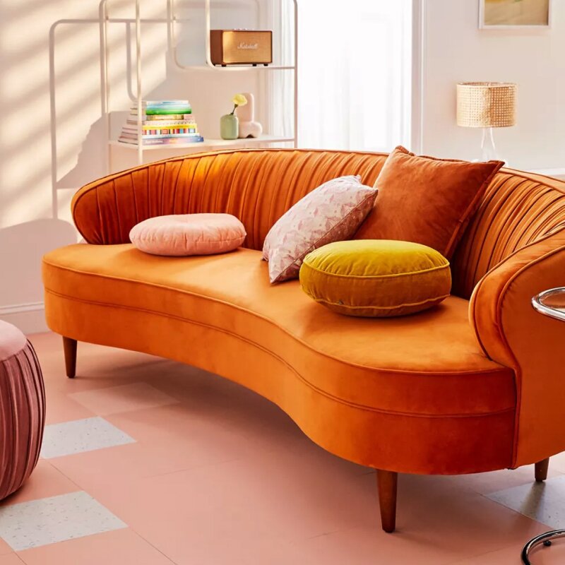 A stylish living room features a vibrant orange velvet curved sofa adorned with three decorative pillows in pink, mustard, and burnt orange. The room is decorated with bookshelves, a small wall-mounted radio, and a lamp on a round side table. The floor is a mix of pink and white tiles.