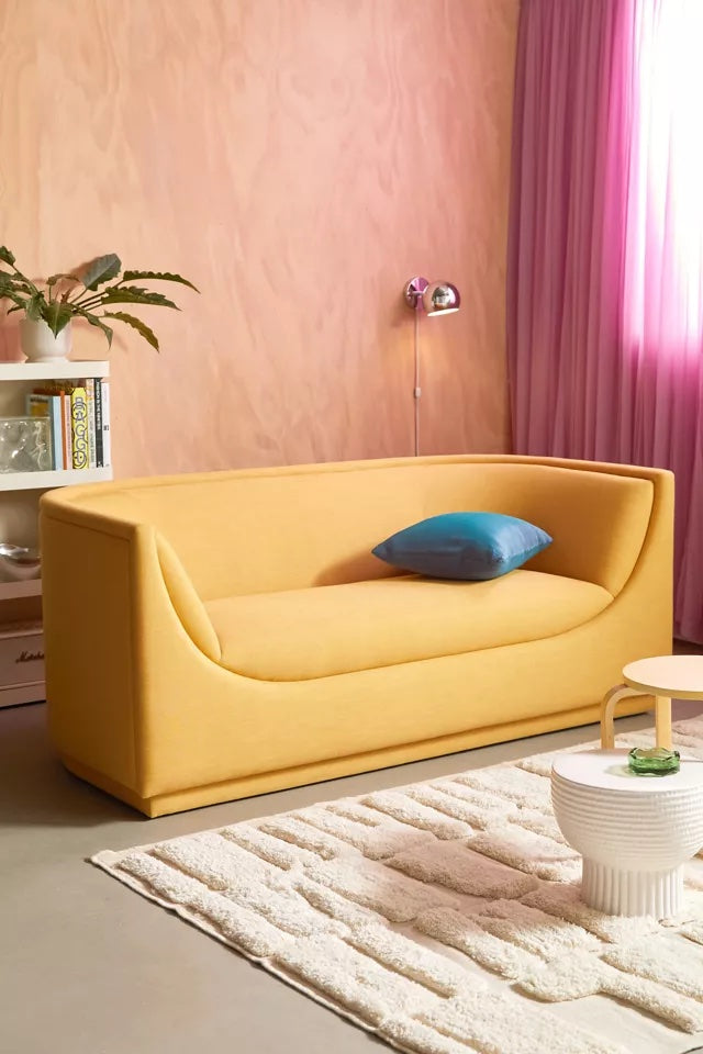 A modern living room features a mustard yellow curved sofa with a blue throw pillow. A white textured rug lies in front of the sofa, and a small round table with a decorative item is placed on the rug. A standing lamp and shelf are nearby, with a pink curtain by the window.