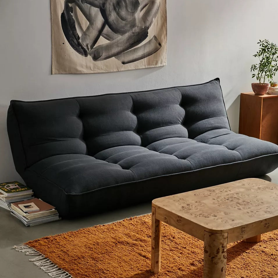 A modern living room featuring a plush black futon with tufted details against a neutral wall. An abstract black and white artwork hangs above the futon. A light wooden coffee table sits on a textured orange rug, with potted plants and stacked magazines beside it.
