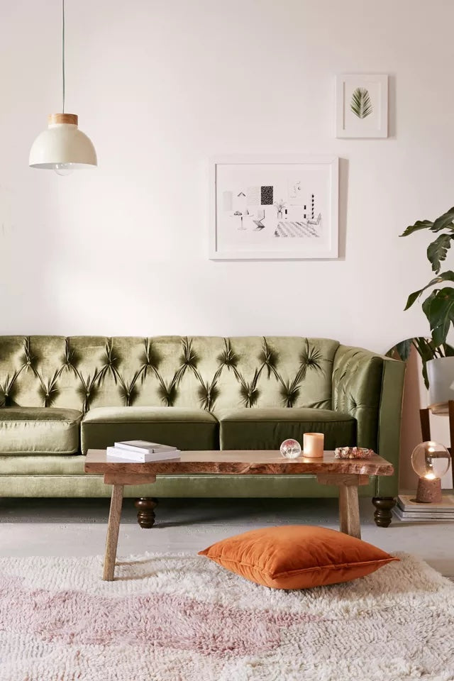 A stylish living room with a green velvet tufted sofa, a wooden coffee table, and an orange floor cushion. The wall behind the sofa features two framed artworks, a hanging pendant light, and a potted plant in the corner. The floor has a plush white rug.
