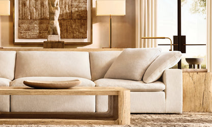 A cozy living room featuring a Cloud Modular 3-Piece Sofa in cream, accompanied by a wooden coffee table and a decorative bowl. Behind the sofa, there is a large textured artwork along with two wall-mounted lamps. Soft lighting and neutral tones enhance the warm atmosphere.